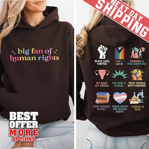 Pode incluir: Um moletom marrom com a frase "big fan of human rights" em letras coloridas na frente. A parte de trás apresenta vários símbolos e frases de justiça social, incluindo "Black Lives Matter" e "Love is Love".