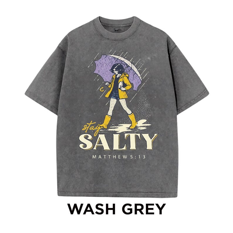 May include: Wash grey t-shirt with a graphic of a person walking in the rain under an umbrella. The design includes the words "stay SALTY" and "MATTHEW 5:13". The person is wearing a yellow jacket and boots.