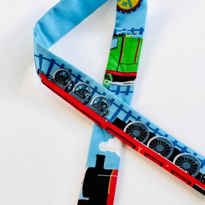 May include: A blue and red fabric strap with a train theme. The strap features a black, red, and green train with blue wheels and a blue background with white clouds. The strap also features a yellow circle with a black cross.