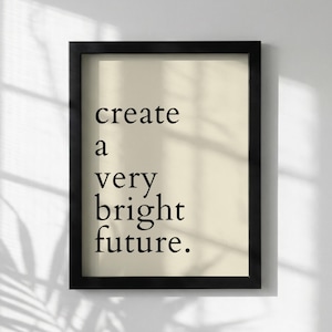 May include: Framed art print with the words "create a very bright future" in black font on a cream background. The black frame has a simple, modern design. The print is displayed against a white wall with shadows.