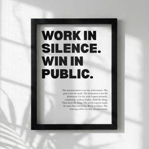 May include: Black framed print with the text "WORK IN SILENCE. WIN IN PUBLIC." in bold, sans-serif font. Below the main text is a paragraph of smaller text. The print is on a white background.