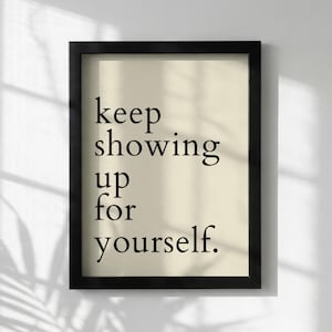 Könnte beinhalten: Ein gerahmter Druck mit dem Text "keep showing up for yourself." Der Text in schwarzer Schriftart befindet sich auf einem cremefarbenen Hintergrund. Der rechteckige Rahmen ist schwarz. Der Druck wird vor einer weißen Wand mit Schatten gezeigt.