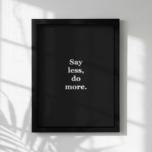 Puede incluir: Una impresión enmarcada en negro con el texto blanco "Say less, do more". La impresión se muestra contra una pared blanca, con sombras de una persiana visible.