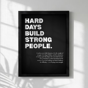 Könnte beinhalten: Schwarz gerahmter Druck mit weißem Text, der lautet: "HARD DAYS BUILD STRONG PEOPLE." Unter dem Haupttext befindet sich ein Textabsatz. Der Druck wird vor einer weißen Wand mit Schatten angezeigt.