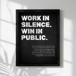 May include: Black framed print with white text that reads "WORK IN SILENCE. WIN IN PUBLIC." Below the main text is a paragraph of text. The print is displayed against a white wall.
