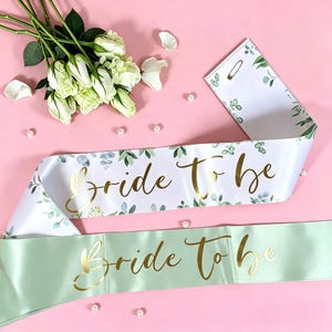 以下が含まれることがあります： 「Bride To Be」のゴールド文字が入ったサテンサッシュ2本。1本は緑の葉のアクセントが付いた白、もう1本は淡い緑色。白いバラと真珠がピンク色の表面に散らばっています。