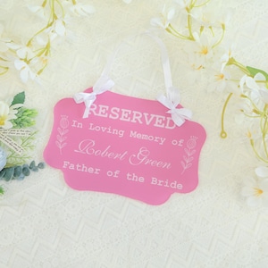May include: A pink sign with white text that reads "RESERVED In Loving Memory of Robert Green Father of the Bride." The sign has a decorative shape and is adorned with white ribbons and floral accents.