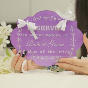 May include: A lavender-colored sign with white text and decorative leaf accents. The sign reads "RESERVED" and "In Loving Memory of Robert Green, Father of the Bride." White ribbons are attached at the top.
