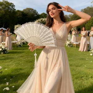 以下が含まれることがあります： 明るいピーチ色のフォーマルドレスを着た女性が、タッセル付きの白いレースの扇子を持っています。扇子は開いており、その複雑なデザインが明らかになっています。同様のドレスを着た他の女性が背景に立っており、結婚式やフォーマルなイベントを示唆しています。