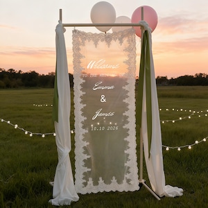 May include: A rectangular wedding welcome sign featuring a sheer white fabric panel with intricate lace borders. The text reads: Welcome TO THE WEDDING OF Emma & James 10.10.2026. The sign is suspended from a metal frame draped with white and olive green fabric.
