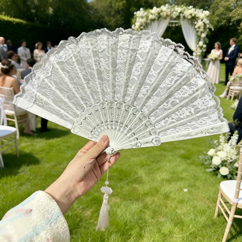 以下が含まれることがあります： 結婚式の前に持たれた、装飾的なタッセルが付いた白いレースの扇子。扇子は複雑なレースのディテールと繊細なデザインが特徴です。背景には結婚式のアーチと緑の芝生にいるゲストが写っています。