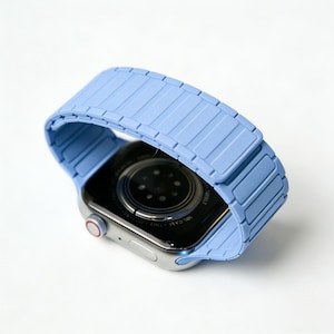 Könnte beinhalten: Ein hellblaues Uhrenarmband ist an einer silbernen Smartwatch befestigt. Das Armband besteht aus einem segmentierten Material. Das Zifferblatt der Uhr ist schwarz mit einem silbernen Rand und einem roten Knopf. Die Uhr ist leicht angewinkelt.