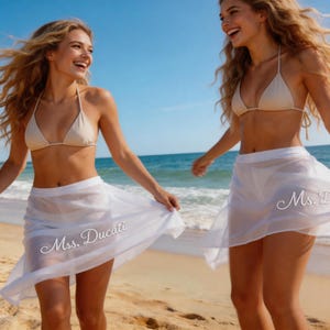 May include: Two women on a beach wearing beige bikini tops and white sheer skirts. The skirts have the text "Ms. Ducati" printed on them. The background shows the sea and a clear blue sky.