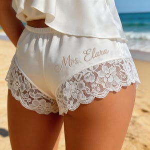 Pode incluir: Calções de cetim marfim com uma delicada barra de renda. Os calções apresentam o texto bordado "Mrs. Elara" em fonte cursiva. Os calções fazem parte de um conjunto de noiva ou lingerie.