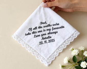 May include: A white handkerchief with a delicate lace trim, held by a hand. The handkerchief features embroidered text in black, including a personal message and a date. The overall design is simple and elegant.