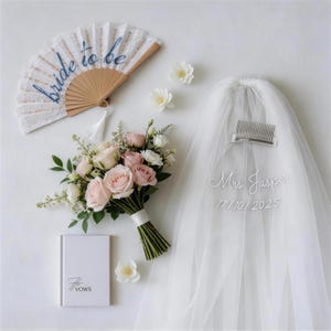 以下が含まれることがあります： 白い扇子に青い文字で「bride to be」、ピンクと白のバラの花束、ベールに「Mrs Jasper 17/10/2025」、そして表紙に「The Vows」と書かれた小さな本が写った、結婚式のフラットレイ。