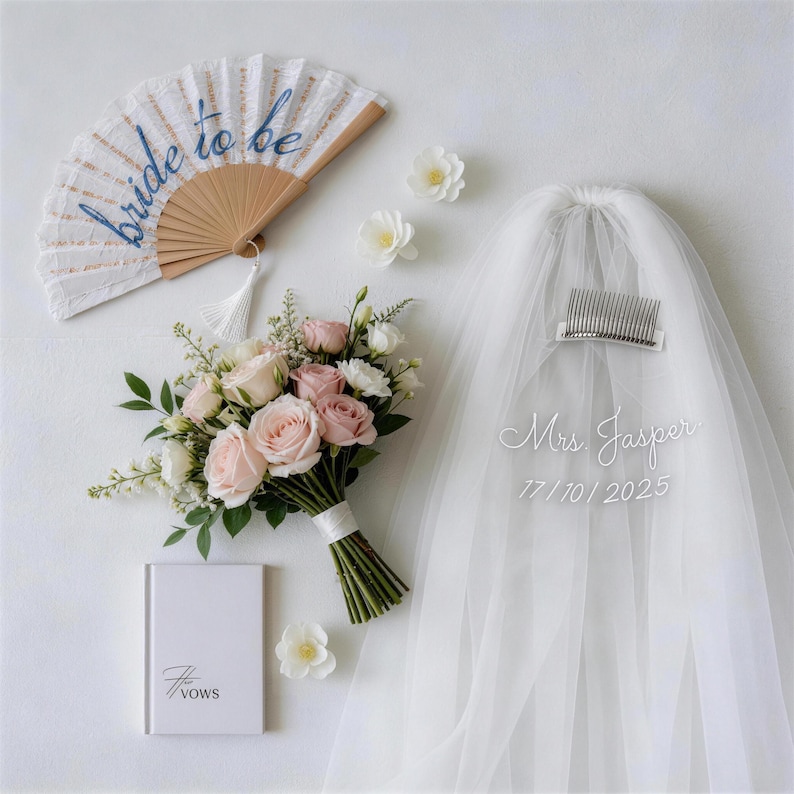 以下が含まれることがあります： 白い扇子に青い文字で「bride to be」、ピンクと白のバラの花束、ベールに「Mrs Jasper 17/10/2025」、そして表紙に「The Vows」と書かれた小さな本が写った、結婚式のフラットレイ。