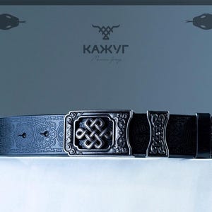 May include: A black leather belt with an ornate silver buckle featuring an intricate knot design. The belt has a repeating pattern and the brand name "KAЖУГ" is visible in the background.