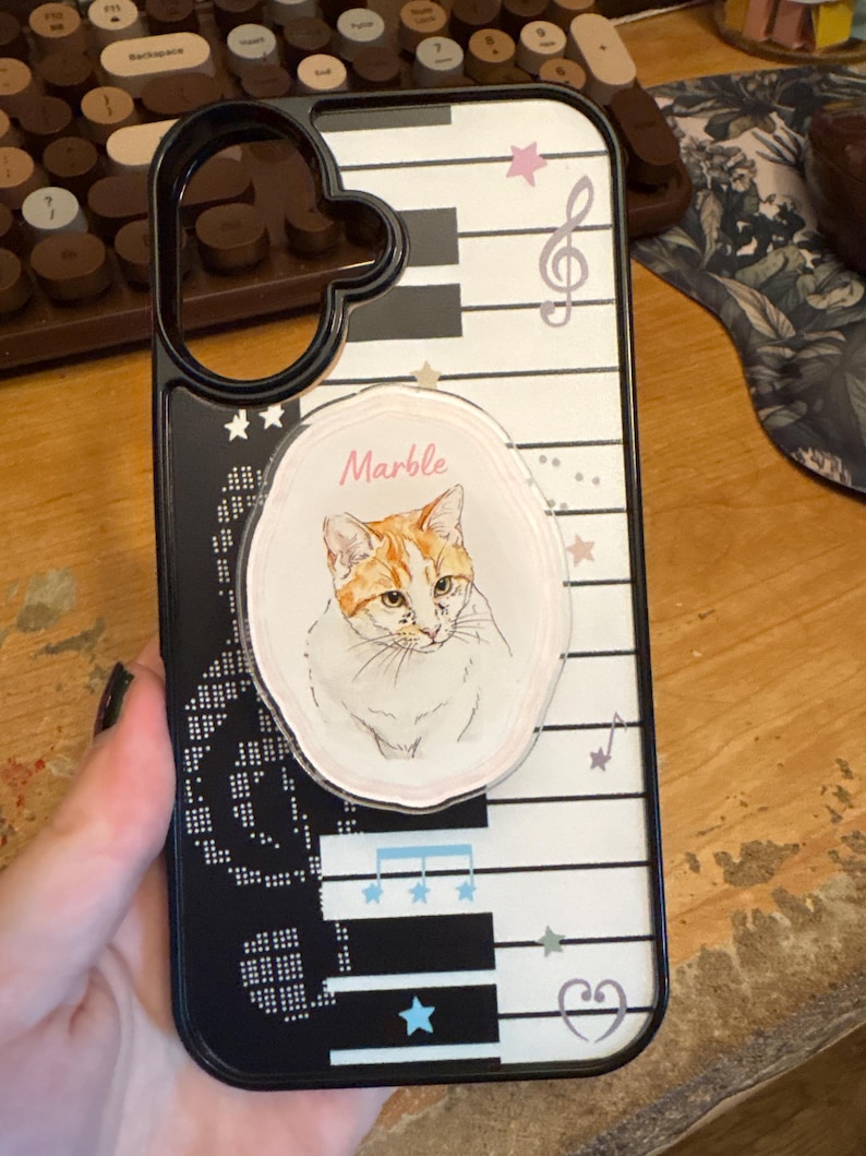 Op de afbeelding: Een zwart-witte telefoonhoes met een pianotoetsenpatroon, muzieknoten en sterren. Een ronde grip is in het midden van de hoes bevestigd en toont een illustratie van een rood-witte kat met het woord Marble erboven geschreven.