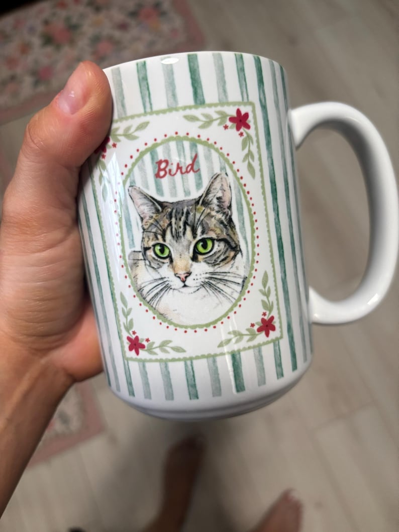 Puede incluir: Una taza de cer&aacute;mica blanca con un patr&oacute;n de rayas verticales verdes y blancas. El centro muestra un marco rectangular con una ilustraci&oacute;n en acuarela de un gato atigrado de ojos verdes, la palabra Bird en rojo y detalles florales.