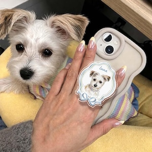 Op de afbeelding: Een beige telefoonhoesje met een ronde, geschulpte acryl gripbevestiging. De grip toont een gedetailleerde illustratie van een kleine witte hond met de naam Luna in blauw schrift erboven en een blauwe strik aan de onderkant.