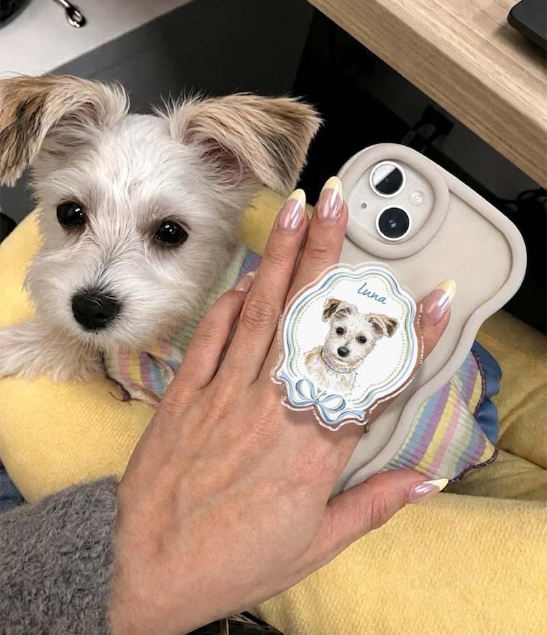 Op de afbeelding: Een beige telefoonhoesje met een ronde, geschulpte acryl gripbevestiging. De grip toont een gedetailleerde illustratie van een kleine witte hond met de naam Luna in blauw schrift erboven en een blauwe strik aan de onderkant.