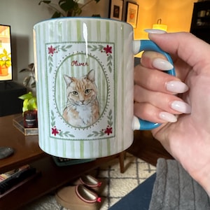 Puede incluir: Una taza de cer&aacute;mica con un patr&oacute;n de rayas verticales de color verde claro y un asa azul. El frente presenta un marco ovalado central que contiene una ilustraci&oacute;n de un gato atigrado naranja con el nombre Oliver escrito encima, con flores rojas.