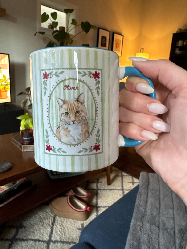 Puede incluir: Una taza de cer&aacute;mica con un patr&oacute;n de rayas verticales de color verde claro y un asa azul. El frente presenta un marco ovalado central que contiene una ilustraci&oacute;n de un gato atigrado naranja con el nombre Oliver escrito encima, con flores rojas.