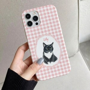 Op de afbeelding: Een telefoonhoesje met een lichtroze en wit ruitpatroon. In het midden bevindt zich een wit ovaal kader met een geschulpte rand, met daarin een illustratie van een zwart-witte kat en de naam Lily in roze schrijfletters boven de kop.