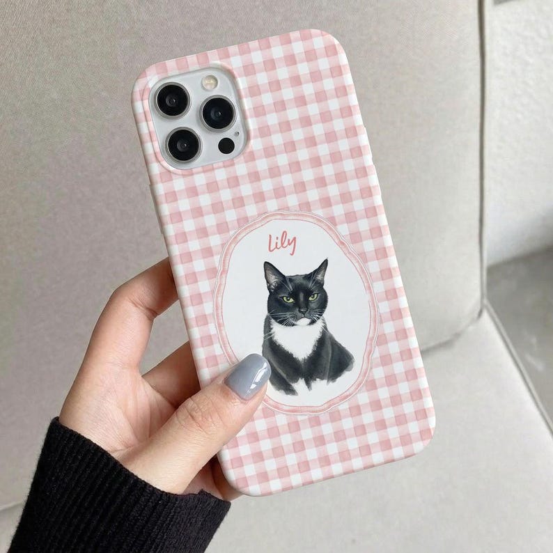 Op de afbeelding: Een telefoonhoesje met een lichtroze en wit ruitpatroon. In het midden bevindt zich een wit ovaal kader met een geschulpte rand, met daarin een illustratie van een zwart-witte kat en de naam Lily in roze schrijfletters boven de kop.