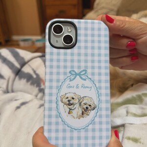 Op de afbeelding: Een lichtblauw en wit telefoonhoesje met een gingham-ruitpatroon en een centraal ovaal kader met een blauwe strik aan de bovenkant. In het kader staat een illustratie van twee kleine, lichtgekleurde honden met de tekst Gus & Remy erboven.