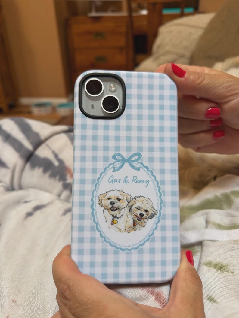 Op de afbeelding: Een lichtblauw en wit telefoonhoesje met een gingham-ruitpatroon en een centraal ovaal kader met een blauwe strik aan de bovenkant. In het kader staat een illustratie van twee kleine, lichtgekleurde honden met de tekst Gus & Remy erboven.