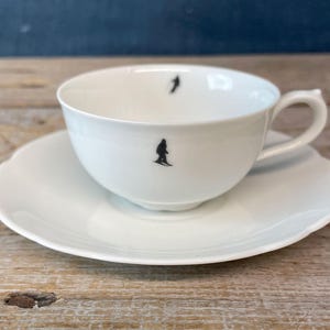 May include: White teacup and saucer set. The teacup has a handle and a black silhouette of a skier. The saucer has a scalloped edge. The set is on a wooden surface.