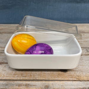 May include: A white rectangular container with a clear lid, holding two decorative eggs. One egg is yellow with orange streaks, and the other is purple. The container sits on a wooden surface.
