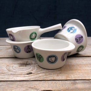 May include: A collection of white ceramic bowls and a small pitcher with handles. Each piece features colorful circular designs with illustrations of vegetables. The bowls are arranged on a wooden surface against a dark blue backdrop.