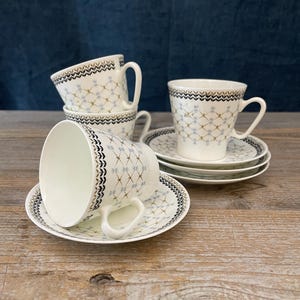 Peut inclure: Un ensemble de tasses à thé et soucoupes en céramique blanche avec un motif répétitif de croix dorées et bleu clair, rehaussé par une bordure en chevron noire et dorée sur le rebord. L'ensemble comprend trois tasses et trois soucoupes assorties.