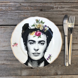 May include: A white ceramic dinner plate with a gold rim features a black and white portrait of Frida Kahlo with flowers in her hair. The plate has a floral design around the edge.