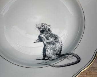 May include: White ceramic plate with a black and white illustration of a rat. The plate has a scalloped edge with a gold trim. The rat is standing on its hind legs, with its tail curled.