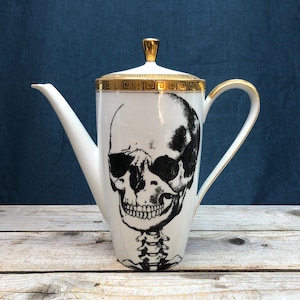 May include: White ceramic teapot with a gold trim and a black and white skull design. The skull has a detailed design with a skeleton underneath.