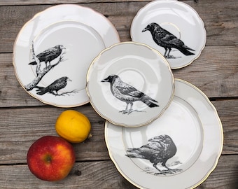 May include: Set of four white ceramic dinner plates with black crow illustrations. The plates have a gold rim and a scalloped edge.