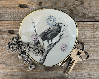 May include: A round white ceramic trinket dish with a gold-tone rim, featuring a black ink illustration of a crow perched on a branch, accented by small blue and pink starburst designs. A silver-tone chain and a metal key rest on the dish.