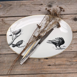 Puede incluir: Plato ovalado blanco con borde dorado e ilustraciones de pájaros negros. Un cuchillo plateado y ramitas de flores secas descansan sobre el plato, colocado sobre una superficie de madera.