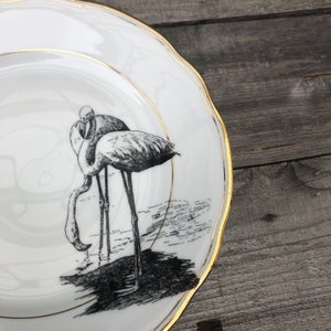 May include: A white ceramic plate with a gold rim features a black and white illustration of two flamingos standing in water.