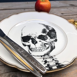 May include: A white plate featuring a black skull design and gold trim. Silver cutlery with decorative patterns is placed on the plate. A red apple is in the background. The plate has a scalloped edge.