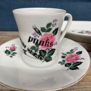 Könnte beinhalten: Weißes Tassen- und Untertassen-Set mit floralem Design und dem Text "punks not dead". Die Tasse und die Untertasse zeigen rosa Rosen, grüne Blätter und lila Akzente. Die Untertasse hat einen Goldrand.