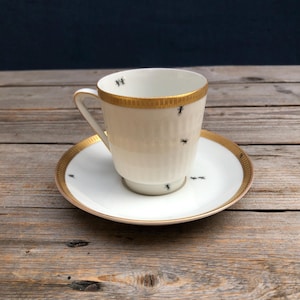 May include: A white porcelain teacup and saucer with a gold rim and black ant design. The cup and saucer are on a wooden surface.