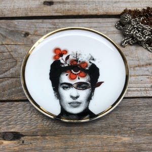 Puede incluir: Un plato de cerámica blanca con borde dorado presenta un retrato en blanco y negro de Frida Kahlo con flores rojas y naranjas en la cabeza.