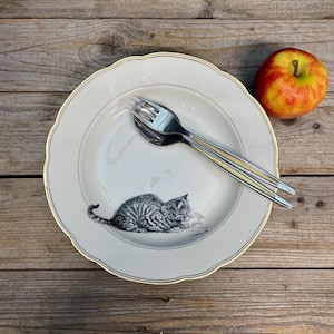 Könnte beinhalten: Ein weißer Keramikteller mit Goldrand und einer schwarz-weißen Illustration einer Katze. Besteck mit goldenen Akzenten liegt auf dem Teller. Ein rot-gelber Apfel liegt daneben. Der Teller steht auf einer Holzoberfläche.