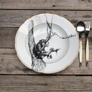 以下が含まれることがあります： 木にいるチンパンジーの白黒イラストが描かれた白いセラミックプレート。プレートは金色の縁とスカラップエッジが付いています。金色の柄の付いたスプーンとフォークが右側にあります。