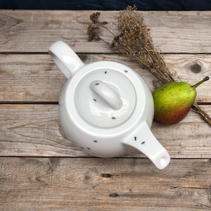 Könnte beinhalten: Weiße Teekanne aus Keramik mit einem schwarzen Ameisenmuster. Die Teekanne hat einen Deckel und einen Ausguss. Die Teekanne steht auf einer Holzoberfläche.